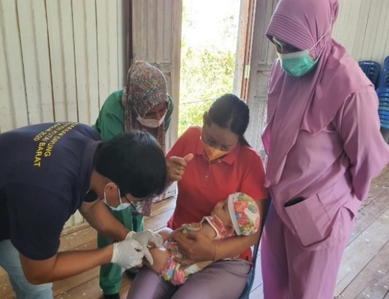 A baby getting immunization while being held by a woman. Three health workers stand around the woman.